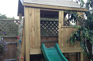 A wooden garden playhouse with a green slide and a climbing wall